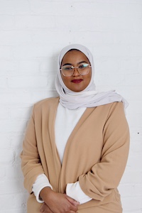 Amani Burale wears glasses and neutral shades of white and beige with her hands folded together casually in front of her.